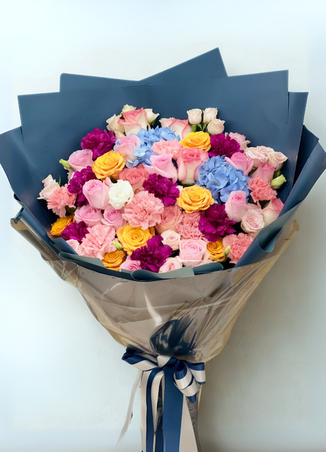 Color Symphony bouquet with pastel roses, blue hydrangeas, and vibrant carnations wrapped in navy | DP FLOWERS BANGKOK
