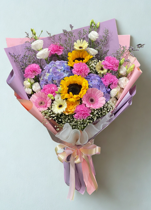 A vibrant mixed flower bouquet with a central sunflower, pink carnations, pink gerbera, and purple hydrangea, wrapped in layered pastel papers - Birthday/Celebration flower delivery Bangkok | DP FLOWERS BANGKOK