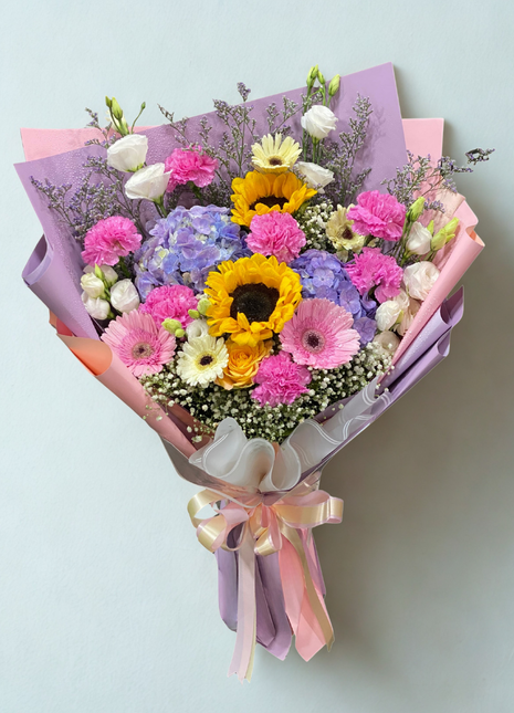 A vibrant mixed flower bouquet with a central sunflower, pink carnations, pink gerbera, and purple hydrangea, wrapped in layered pastel papers - Birthday/Celebration flower delivery Bangkok | DP FLOWERS BANGKOK