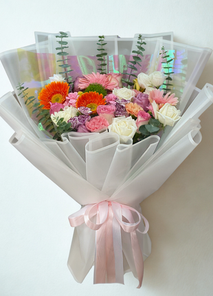 A vibrant mixed flower bouquet with orange gerbera, pink roses, and purple carnations, wrapped in dazzling iridescent/holographic cellophane and white mesh - Trendy/Modern flower delivery Bangkok | DP FLOWERS BANGKOK