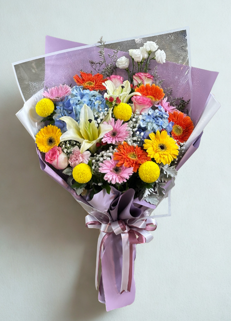 A vibrant mixed flower bouquet with orange, yellow, and pink Gerbera, white Lily, and blue Hydrangea, wrapped in purple and silver paper - Happy Birthday/Celebration flower delivery Bangkok | DP FLOWERS BANGKOK