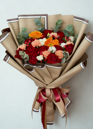 A vibrant bouquet of red roses, orange gerbera, and coral carnations with eucalyptus, wrapped in layered tan/brown paper with red and bronze ribbon - Festive/Celebration flower delivery Bangkok | DP FLOWERS BANGKOK